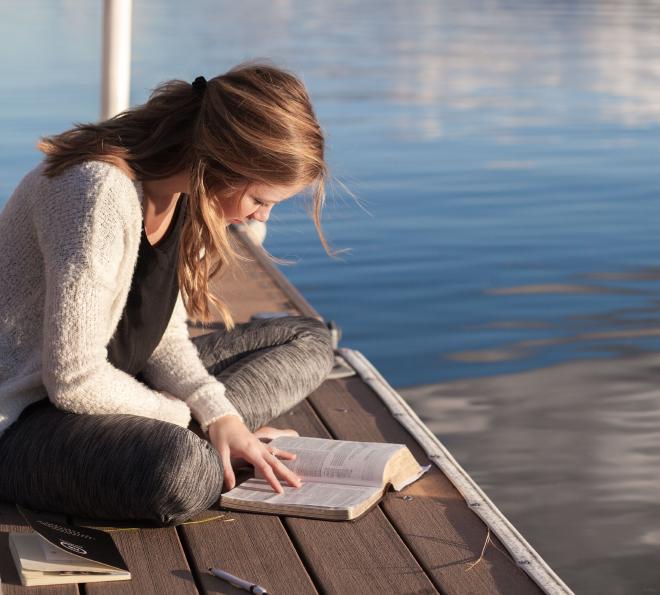 A woman sat down on wooden decking next to a body of water, reading a Bible in contemplation.